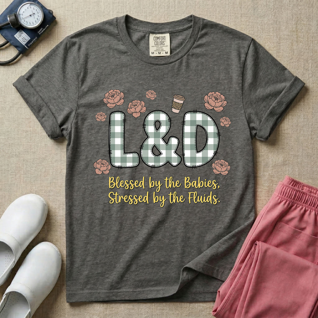 A descriptive graphic tee for labor and delivery nurses featuring plaid block letters spelling L&D surrounded by floral elements and a coffee cup, with the text "Blessed by the Babies, Stressed by the Fluids."