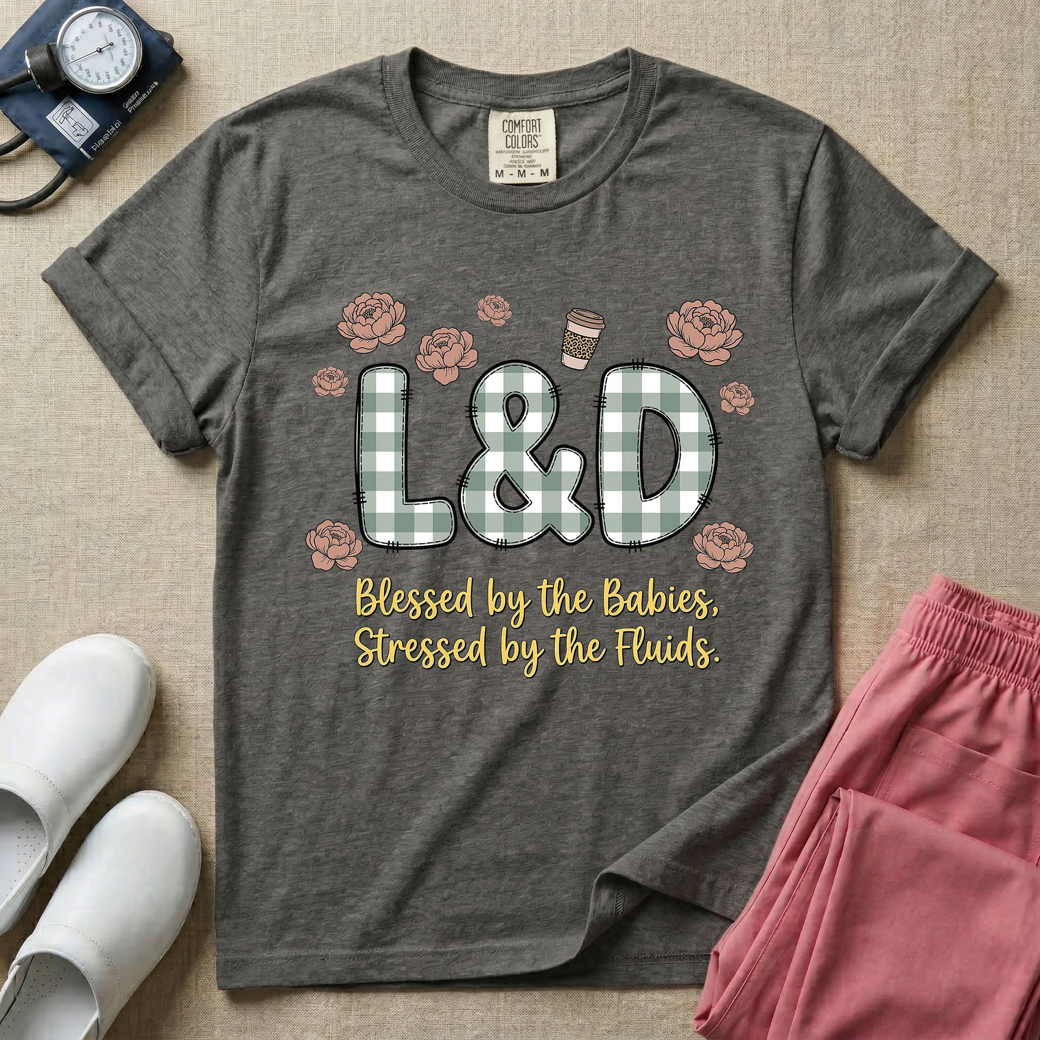 A descriptive graphic tee for labor and delivery nurses featuring plaid block letters spelling L&D surrounded by floral elements and a coffee cup, with the text "Blessed by the Babies, Stressed by the Fluids."