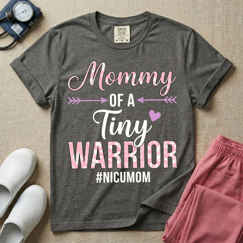 A cute NICU mom t-shirt featuring the stacked text "Mommy OF A Tiny WARRIOR #NICUMOM" in pink and white, accented with decorative arrows, a small pink heart, and a pink plaid pattern filling the word warrior.