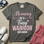 A cute NICU mom t-shirt featuring the stacked text "Mommy OF A Tiny WARRIOR #NICUMOM" in pink and white, accented with decorative arrows, a small pink heart, and a pink plaid pattern filling the word warrior.