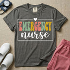 A retro-style t-shirt for emergency room nurses featuring the word "EMERGENCY" in colorful, vintage lettering above the word "nurse" in a delicate white script font, framed by a thin white rectangular border with a small heart at the top center.