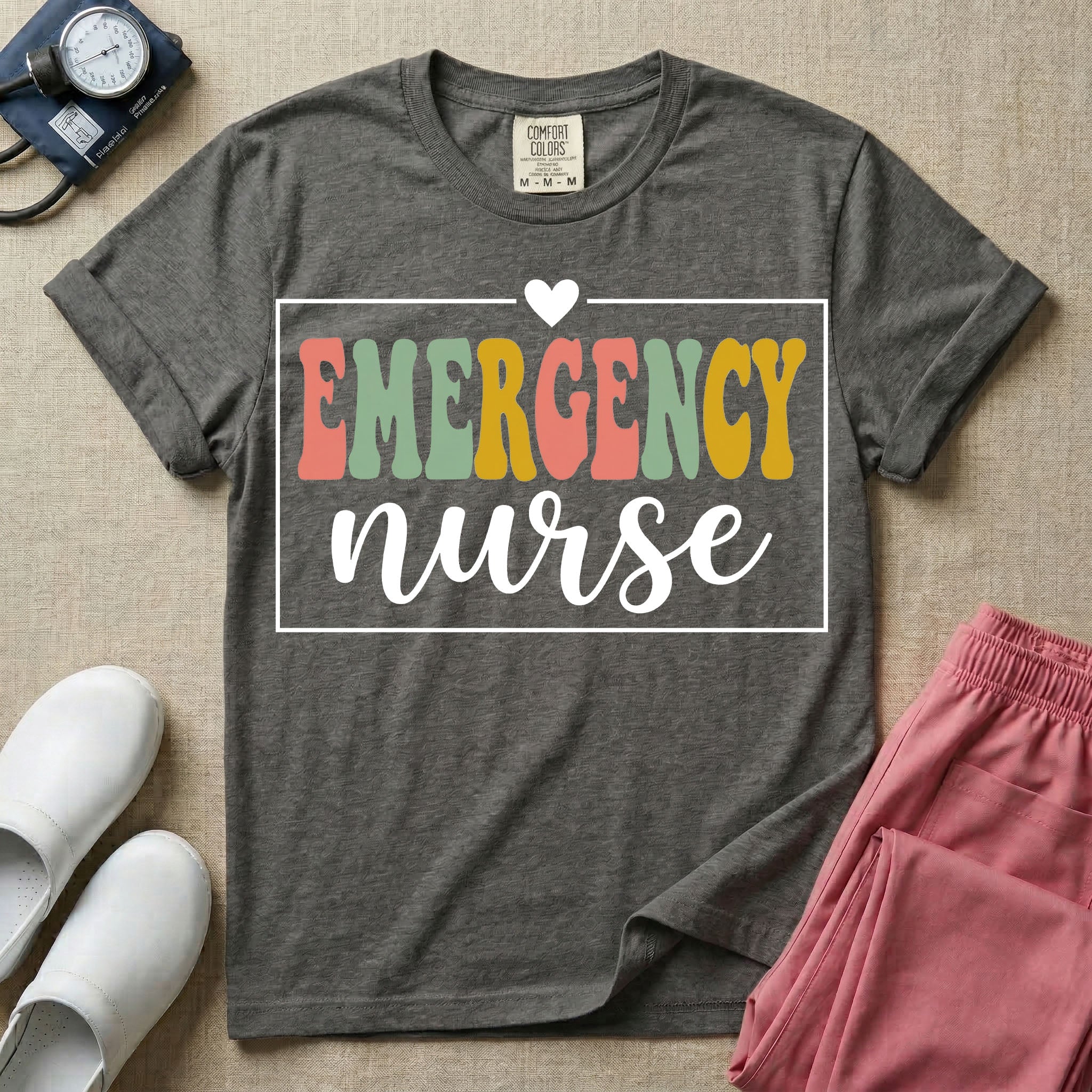 A retro-style t-shirt for emergency room nurses featuring the word "EMERGENCY" in colorful, vintage lettering above the word "nurse" in a delicate white script font, framed by a thin white rectangular border with a small heart at the top center.