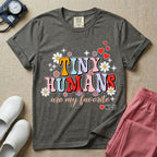 A t-shirt featuring the text Tiny Humans are my favorite in colorful retro bubble letters decorated with daisies, sparkles, and red hearts with a small NICU heart logo at the bottom.