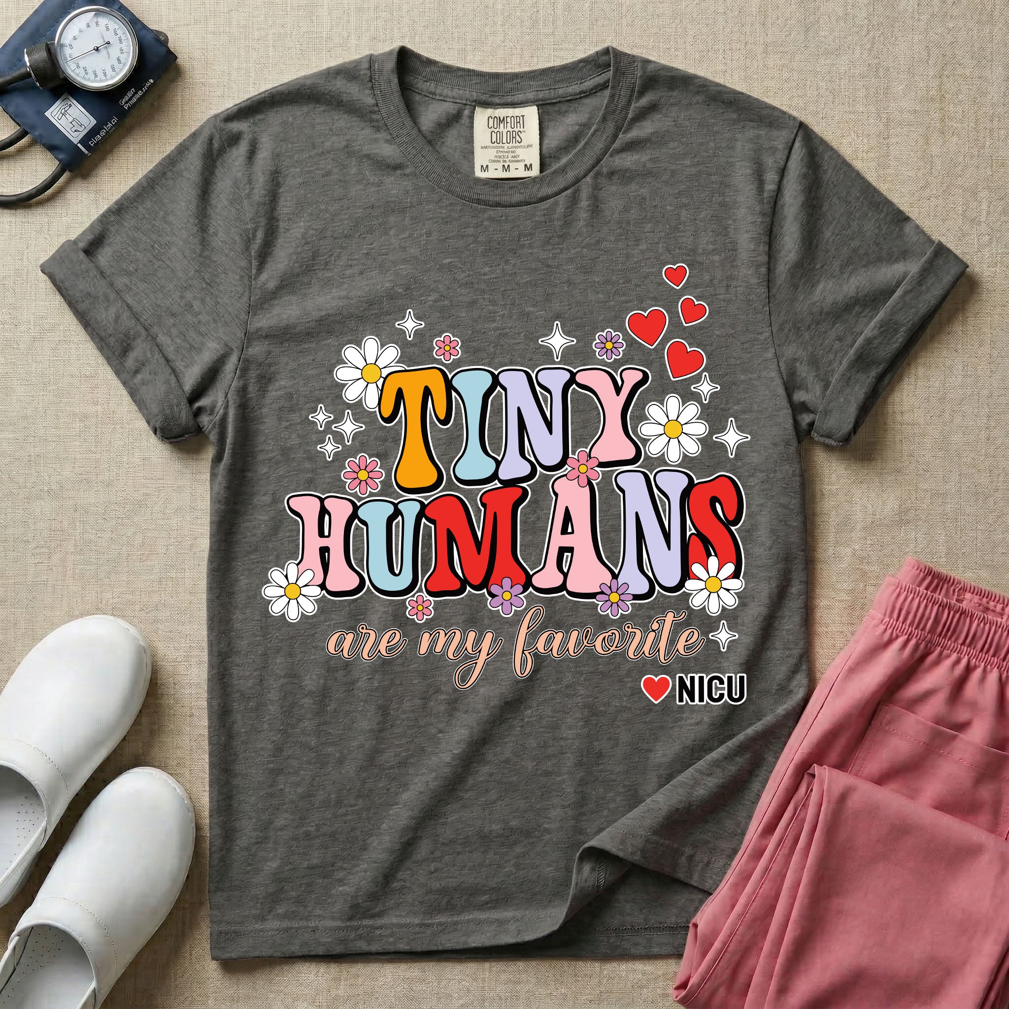 A t-shirt featuring the text Tiny Humans are my favorite in colorful retro bubble letters decorated with daisies, sparkles, and red hearts with a small NICU heart logo at the bottom.