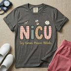 A humorous t-shirt for NICU nurses featuring the large text "NICU" in plaid and patterned letters with the phrase "Tiny Humans. Massive Attitudes." underneath, accompanied by small cute graphics of a syringe, an iced coffee cup, and a baby's face.